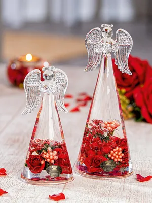 Two glass angel figurines filled with red roses and small white berries, placed on a wooden surface with scattered red rose petals. A lit candle in a red holder is visible in the background.