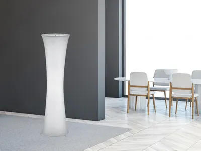 A tall, white, hourglass-shaped floor lamp stands on a light gray rug in a modern room with dark gray walls and a dining area with white chairs and tables visible through an opening.