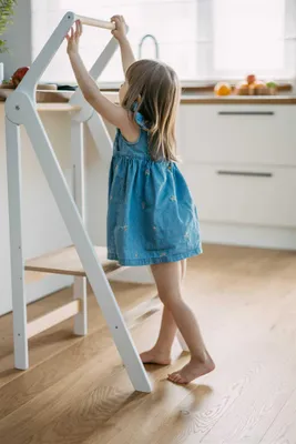 A young girl stands on a wooden floor, reaching up to a white learning tower in a kitchen setting.