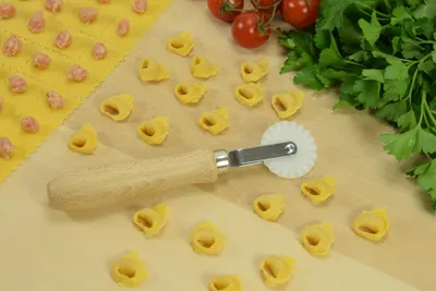 A pasta cutter and tortellini on a wooden surface, with tomatoes and parsley in the background.