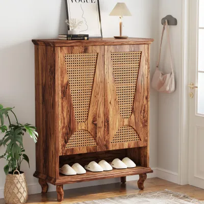 A wooden shoe cabinet with rattan doors is shown in a hallway, with shoes on the bottom shelf and a plant and lamp on top.