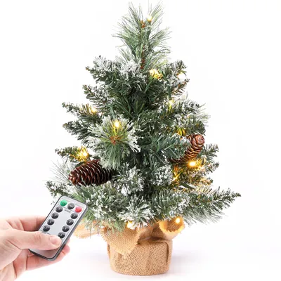 A hand holding a remote control is shown in front of a small, decorated Christmas tree.