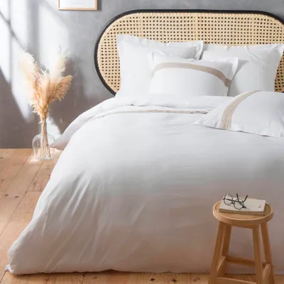 A neatly made bed with white bedding and decorative pillows, set against a grey wall and wooden floor. A vase of dried pampas grass is on the left, and a small wooden stool with glasses and a book is on the right.