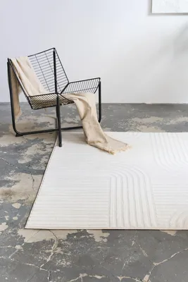 A modern chair with a beige throw draped over it sits next to a white rug with a geometric pattern on a concrete floor.
