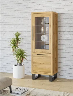 A wooden display cabinet with glass doors and a drawer, placed in a room with a white brick wall and a potted plant.