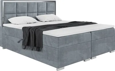 A studio shot of a gray upholstered box spring bed with a headboard, two pillows, and a dark gray throw blanket.