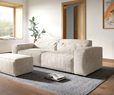 A modern living room with a beige sofa and ottoman on a rug, next to a window and a doorway.