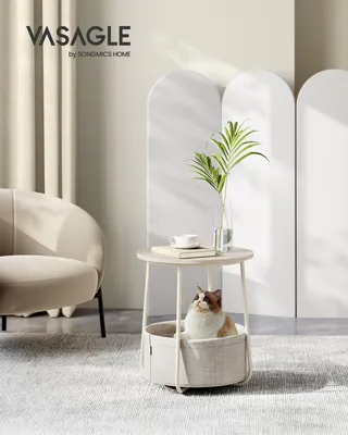 A cat sits in a round basket-style side table, with a chair and decorative screens in the background.