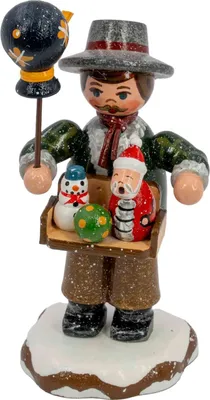Ein handgefertigter Holzfigur zeigt einen Spielwarenhändler mit einem Tablett voller bunter Spielzeuge in den Händen und einem Vogel auf einem Stock während er auf einem schneebedeckten Sockel steht