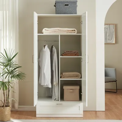 An open white wardrobe with clothes and folded blankets inside, placed in a room with a plant and a chair visible in the background.