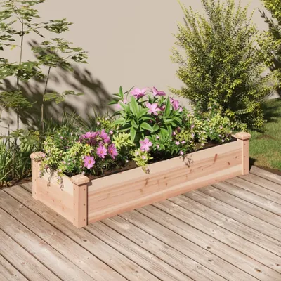A wooden planter box filled with pink and green plants sits on a wooden deck, with a wall and bushes in the background.