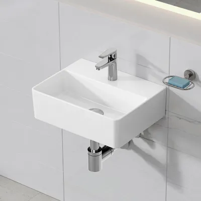 A white ceramic wall-mounted sink with a chrome faucet and drainpipe, mounted on a white tiled wall. A silver soap dish with a blue bar of soap is attached to the wall to the right of the sink.