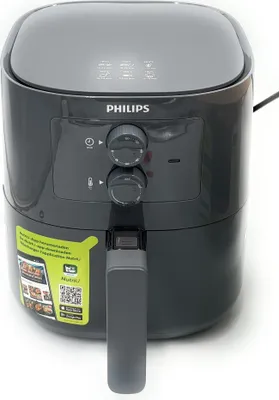 A dark gray Philips air fryer is shown against a white background. The air fryer has a digital display on top and two knobs on the front for controls. A yellow sticker with text and a QR code is attached to the side of the air fryer.