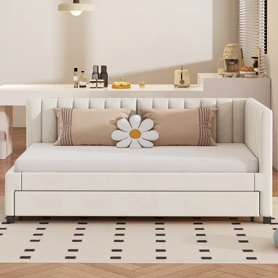 A beige upholstered daybed with a pull-out trundle bed, shown in a bright, minimalist room setting with decorative pillows and a rug.
