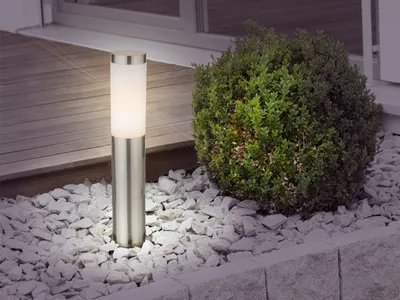 A modern outdoor lamp stands in a garden bed with gravel and a bush.
