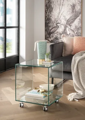 A glass side table with books and bowls on it, next to a sofa with cushions and a blanket.