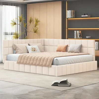 A modern bedroom with a beige upholstered bed, pillows, and a blanket. A plant is visible in the background, along with shelves of books.