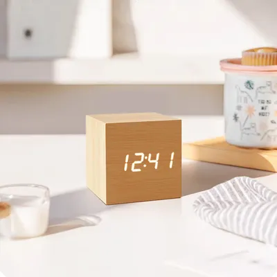 A wooden cube digital clock displaying the time 12:41 is placed on a white surface next to a glass and a striped cloth. In the background, a white shelf and a decorative cup are visible.