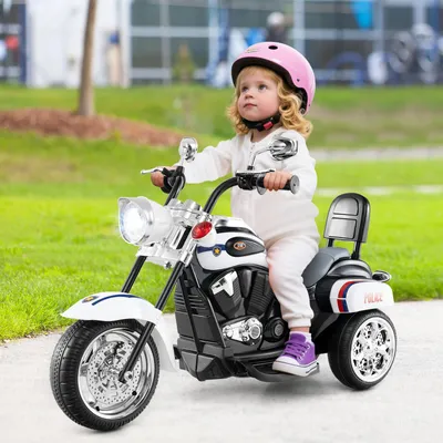 A young child rides a toy police motorcycle outdoors on a paved path, with grass and trees in the background.