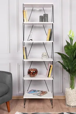 A tall, narrow white bookshelf with black metal legs is displayed against a white paneled wall. Decorative items like books, candles, and a glass bowl are placed on its shelves. A gray armchair and a large potted plant are visible to the left and right, re