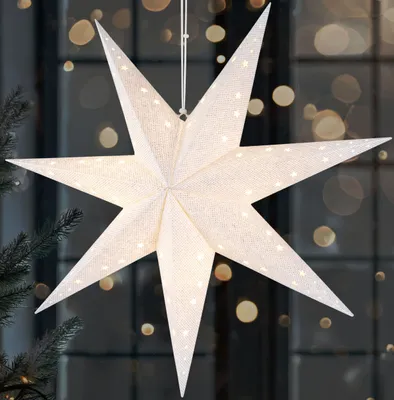 A white, star-shaped paper lantern with small star cutouts is illuminated from within, hanging in front of a window with blurred lights.
