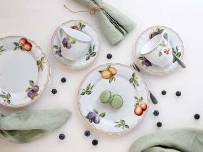 A top-down view of a fruit-themed porcelain tea set with two cups and saucers, three plates, and two macarons, arranged on a white background with scattered blueberries and green napkins.