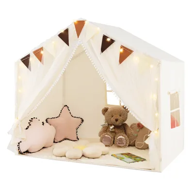 A children's play tent shaped like a house, decorated with bunting and fairy lights, and furnished with pillows, a teddy bear, and a gingerbread man figure.