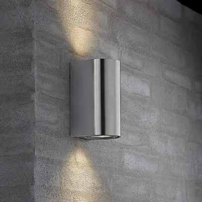A modern, cylindrical outdoor wall light made of stainless steel is mounted on a textured gray brick wall. The light is illuminated, casting upward and downward beams of warm light.