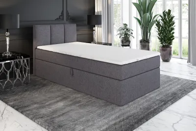 A bedroom scene featuring a gray boxspring bed with a white mattress and topper, set on a gray rug.
