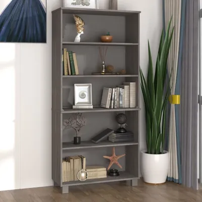 A gray wooden bookshelf filled with books and decorative items, placed in a room with a large plant and curtains.