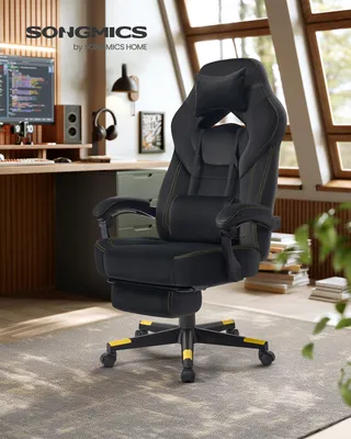 A black gaming chair with yellow accents is shown in a home office setting with a desk, computer, and window in the background.