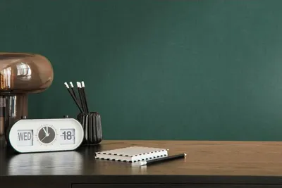 A desk with a clock, notebook, pencils, and a lamp against a dark green wall.