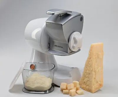 A white and silver electric cheese grater with a block of cheese, grated cheese in a container, and cubed cheese.