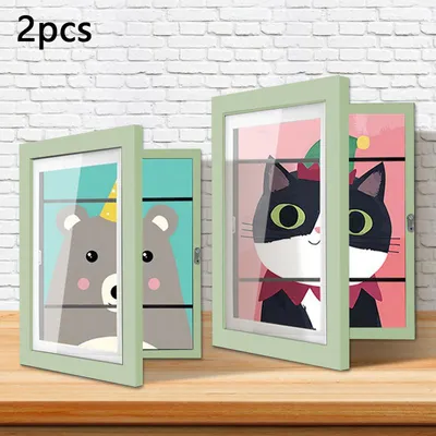 Two open picture frames displaying cartoon animal art are placed on a wooden surface against a white brick wall.