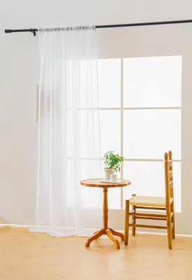 A sheer white curtain hangs on a curtain rod in front of a large window. A small wooden table with a potted plant and a wooden chair are placed in front of the window.