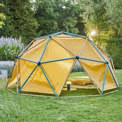 A yellow geodesic dome tent structure with blue and brown poles is set up on a green lawn in a backyard. Pillows and a small lamp are inside the tent.
