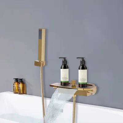 A gold waterfall faucet with a hand shower attachment is mounted on a gray wall above a white bathtub. Two black pump bottles of toiletries are on a shelf attached to the faucet.