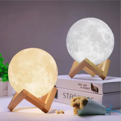Two moon lamps on wooden stands, one lit with a warm yellow light and the other with a cool white light, placed on top of books.
