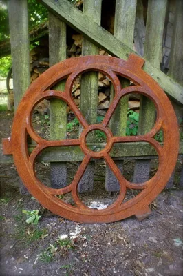 A rusty metal decorative window frame leans against a wooden fence with a pile of firewood in the background.