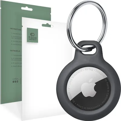 A dark gray protective case for an Apple AirTag is shown, attached to a keychain ring. Next to it are two white packaging inserts, one with a green logo and text, and another with green text on a green background.