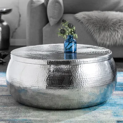 A round, hammered metal coffee table with a blue vase and flowers on top, placed on a rug in a living room setting.