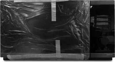 A black Samsung microwave oven is covered in plastic wrap and secured with tape.