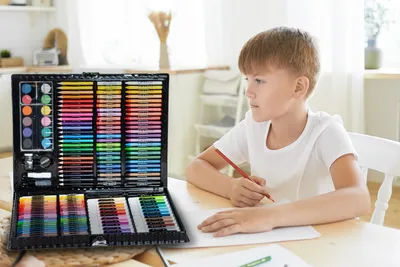 A young boy is drawing at a table with an open art set in front of him.