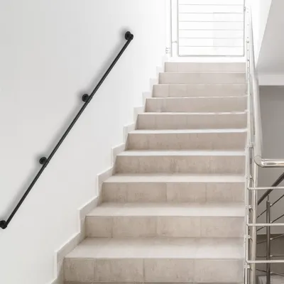 A black metal handrail is mounted on a white wall next to a light-colored stone staircase. The staircase leads upwards, with a metal railing on the right side.