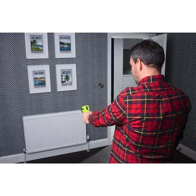 A man in a red plaid shirt is using a yellow and black infrared thermometer to measure the temperature of a white radiator in a room with patterned wallpaper.
