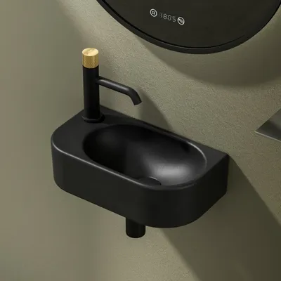 A black wall-mounted sink with a black faucet and a gold accent on the faucet handle, set against a textured greenish-gray wall. A portion of a circular mirror with digital display is visible in the upper background.
