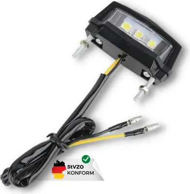 A close-up shot of a black license plate light with LED bulbs, featuring attached wires and a German conformity sticker.