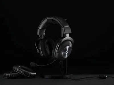 A black Logitech G PRO X gaming headset is displayed on a black stand against a black background. Two extra ear pads are placed in the foreground.