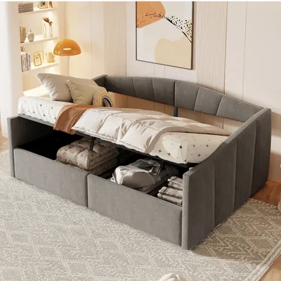 A grey upholstered daybed with storage compartments open, revealing folded blankets and linens inside. The daybed is set up in a room with a patterned rug and a framed artwork on the wall.