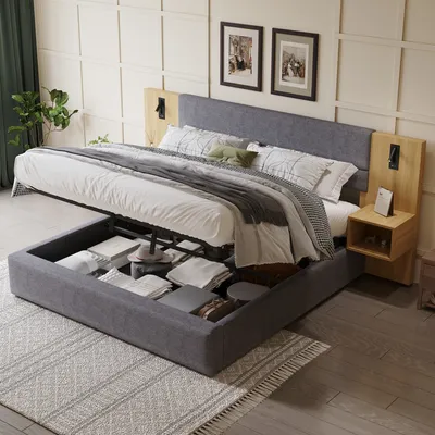 A bedroom scene featuring a large storage bed with the mattress lifted to reveal storage space underneath. The bed is made with white and gray bedding. Two framed pictures hang on the wall above the headboard, and a small wooden nightstand with a lamp is a